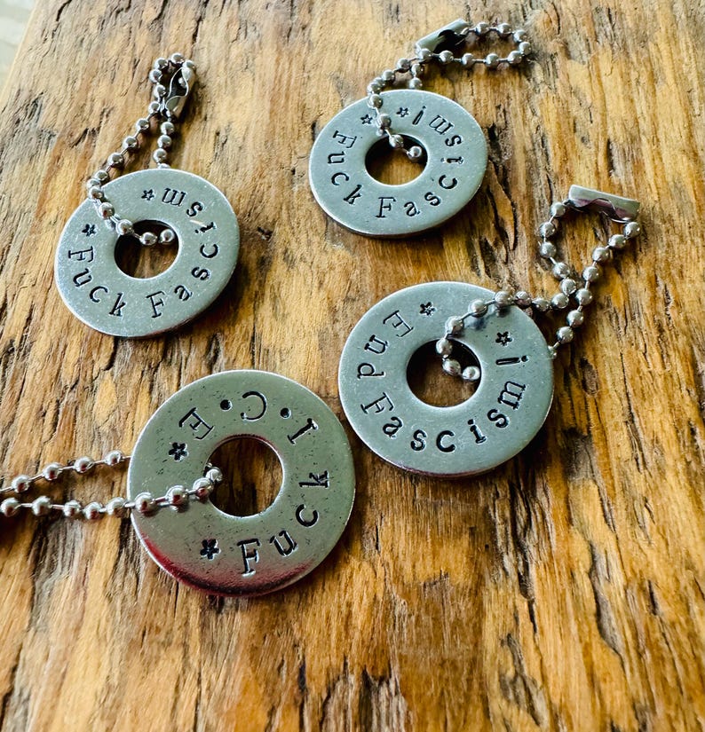 Puede incluir: Cuatro colgantes plateados con cadenas de bolas. Cada colgante es un c&iacute;rculo plano con un agujero en el centro y texto estampado que dice "Fuck Fascism" o "End Fascism!". Los colgantes se muestran sobre una superficie de madera.