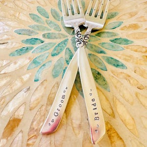 May include: Two silver forks with ornate handles, crossed and engraved with the words "Bride" and "Groom". The forks are on a decorative, iridescent surface.