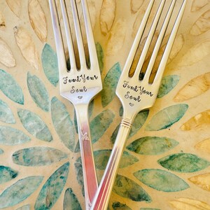 May include: Two silver forks, each engraved with "Feed Your Soul", are crossed over a decorative surface. The surface features a floral pattern in green, blue, and beige hues, creating a contrasting backdrop.