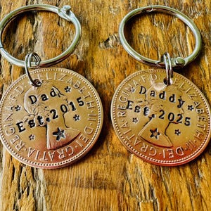 May include: Two circular metal keychains made from coins, each featuring a silver split ring. The left coin is stamped with "Dad" and "Est. 2015," while the right coin is stamped with "Daddy" and "Est. 2025." Both coins feature a profile portrait.