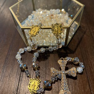 May include: A rosary with clear and gray beads, gold-colored accents, and a decorative cross. The rosary is displayed on a dark wooden surface, with a glass and gold-colored hexagonal box in the background filled with clear beads.