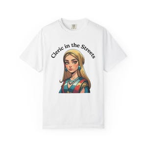 Puede incluir: Camiseta blanca con una ilustración de dibujos animados de una mujer con cabello rubio y pendientes azules. El texto "Cleric in the Streets" está arqueado sobre la ilustración en negro. Prenda de vestir informal.