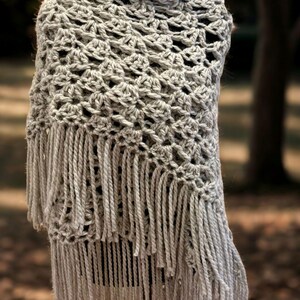 May include: A light gray crochet shawl featuring an intricate openwork pattern of repeating shell stitches. The garment is finished with a long, thick fringe of matching yarn strands hanging from the bottom edge, creating a textured appearance.