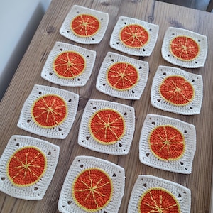 Könnte beinhalten: Eine Sammlung quadratischer Häkeluntersetzer. Jeder Untersetzer hat einen weißen Rand, einen gelben Ring und ein oranges Zitrusfrucht-Design in der Mitte. Die Untersetzer sind auf einer Holzoberfläche angeordnet.