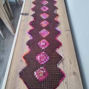 Può includere: Runner da tavolo all'uncinetto marrone con bordo a zig-zag. Presenta motivi floreali quadrati rosa, arancioni e viola. I bordi hanno un bordo rosa. Un elemento decorativo per la casa, lungo circa 150 cm.