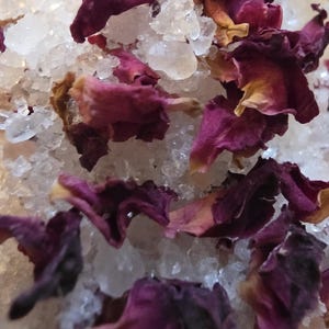 May include: A mixture of coarse, translucent white salt crystals combined with dried, deep magenta and dark pink rose petals. The texture is granular and uneven, featuring a blend of jagged mineral pieces and soft, curled floral elements.