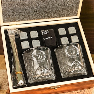 Op de afbeelding: Houten geschenkdoos met twee gegraveerde whiskyglazen, ijsstenen en een tang. De glazen zijn geëtst met de nummers "30" en "18" samen met namen en data. Een fles wijn en een whiskyglas zijn ook zichtbaar.