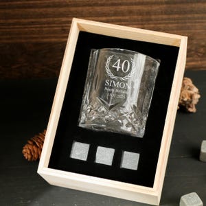 May include: A clear, engraved whiskey glass with the number 40, the name "SIMON", and the date 15.11.2025, presented in a wooden box with three square ice stones. The glass has a decorative pattern.