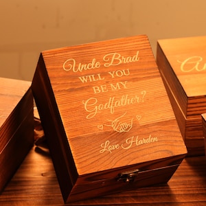Könnte beinhalten: Eine Holzkiste mit dem eingravierten Text "Uncle Brad, WILL YOU BE MY Godfather?" in einer Schreibschrift. Die Kiste ist braun mit einem Metallverschluss und wahrscheinlich eine Geschenkbox.