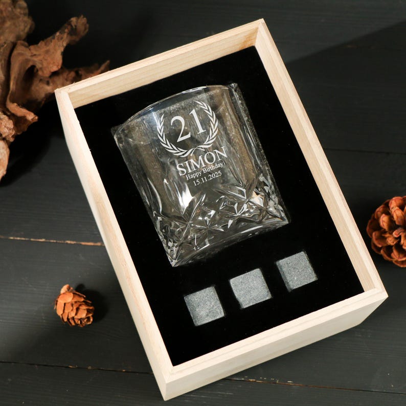 May include: A personalized whiskey glass with the number 21, a laurel wreath, the name "SIMON", and the date "15.11.2025" engraved on it. The glass is in a wooden box with three gray square stones. The box has a black velvet interior.