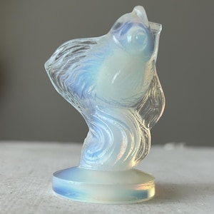 May include: A translucent, milky white opalite figurine shaped like a goldfish with a flowing tail and fins. The sculpture features a smooth, polished surface that catches the light, creating soft blue and iridescent highlights across its form.