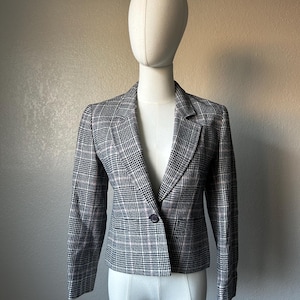 Puede incluir: Blazer a cuadros en negro, blanco y rosa, exhibido en un maniquí. El blazer tiene un cierre de un solo botón y una solapa clásica con muescas. La tela tiene un patrón sutil y las mangas son largas.