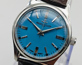 May include: A silver-toned Jaeger-LeCoultre watch with a vibrant blue face and a dark brown leather strap. The watch features silver hands and markers, and a black leather band with white stitching. The watch is displayed on a clear stand.