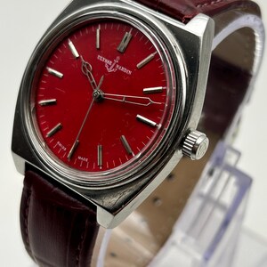 May include: A silver-tone wristwatch with a vibrant red dial featuring the Ulysse Nardin logo and anchor symbol. The watch has silver hour markers, thin silver hands, and a textured dark red leather strap with a silver crown on the side.