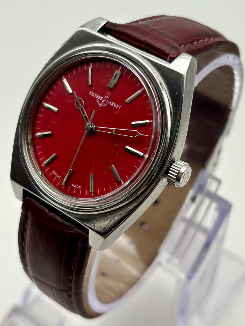 May include: A silver-tone wristwatch with a vibrant red dial featuring the Ulysse Nardin logo and anchor symbol. The watch has silver hour markers, thin silver hands, and a textured dark red leather strap with a silver crown on the side.