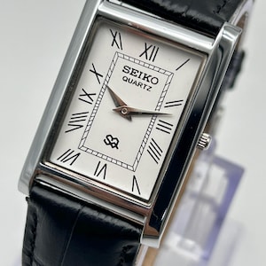 May include: A rectangular Seiko Quartz watch with a white face, black Roman numerals, and silver hands. The watch has a black leather band with a crocodile pattern and a silver case. The watch face displays the Seiko Quartz logo.