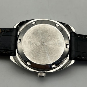 May include: Close-up of the back of a silver-toned wristwatch with a black leather band. The watch face is circular with a brushed metal finish. The band is black with white stitching.