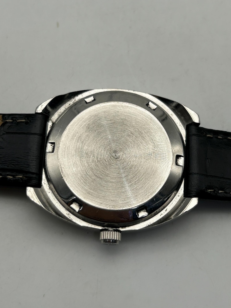 May include: Close-up of the back of a silver-toned wristwatch with a black leather band. The watch face is circular with a brushed metal finish. The band is black with white stitching.
