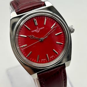 May include: A wristwatch with a circular red dial, silver-tone hour markers, and silver hands. The watch features a silver-tone metal case and a dark red leather strap with a textured finish. The dial displays the text ULYSSE NARDIN and SWISS MADE.