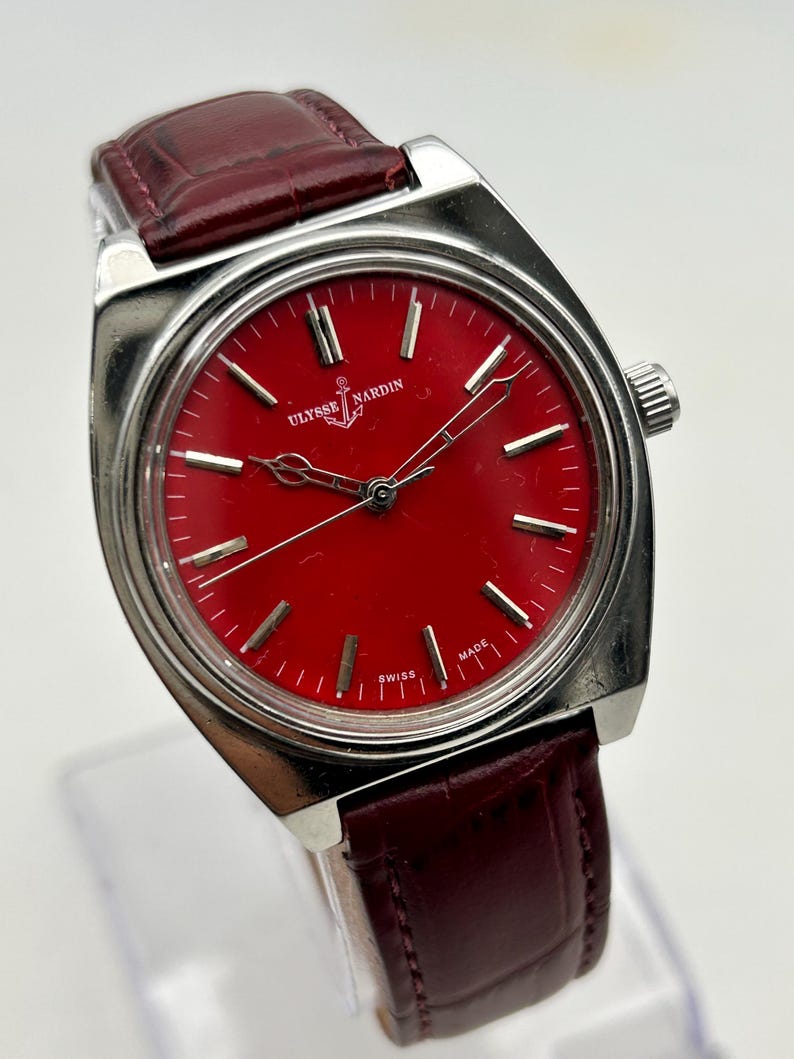May include: A wristwatch with a circular red dial, silver-tone hour markers, and silver hands. The watch features a silver-tone metal case and a dark red leather strap with a textured finish. The dial displays the text ULYSSE NARDIN and SWISS MADE.