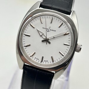 May include: A silver-toned Ulysse Nardin wristwatch with a white face and black leather band. The watch face features the brand name and the words "SWISS MADE". The watch has a classic design with a round face and a black leather strap.