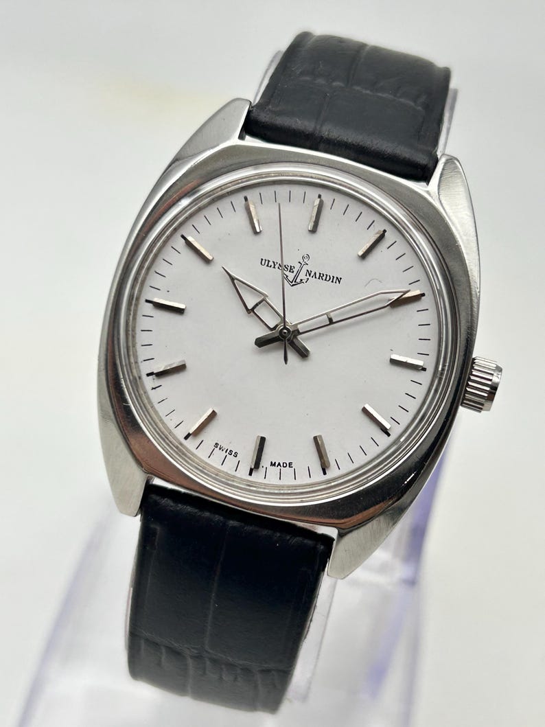 May include: A silver-toned Ulysse Nardin wristwatch with a white face and black leather band. The watch face features the brand name and the words "SWISS MADE". The watch has a classic design with a round face and a black leather strap.