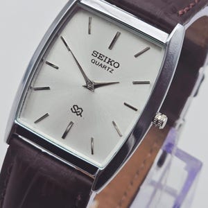Könnte beinhalten: Eine silberne Seiko Quartz-Uhr mit rechteckigem Zifferblatt und braunem Lederarmband. Das Zifferblatt ist silberfarben mit schwarzen Markierungen und Zeigern. Das Armband ist dunkelbraun mit einem strukturierten Krokodilmuster.