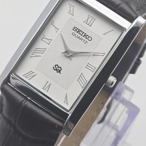 May include: A rectangular Seiko Quartz watch with a silver case and a white face featuring Roman numerals and a textured centre. The watch has a black leather strap with a crocodile pattern.