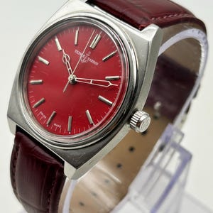 May include: A Ulysse Nardin wristwatch featuring a round, vibrant red dial with silver-toned hour markers and hands. The watch has a polished silver-tone metal case and a dark red, textured leather strap with visible stitching.