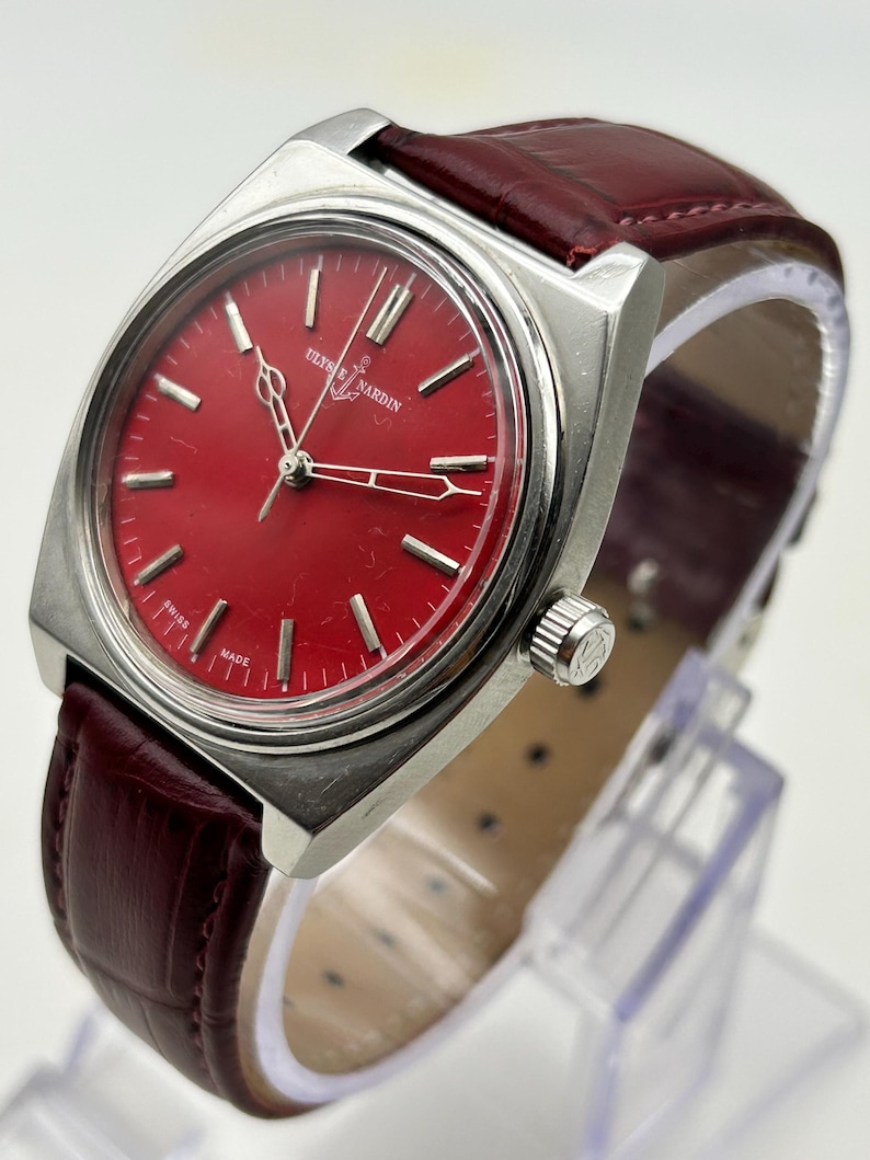May include: A Ulysse Nardin wristwatch featuring a round, vibrant red dial with silver-toned hour markers and hands. The watch has a polished silver-tone metal case and a dark red, textured leather strap with visible stitching.