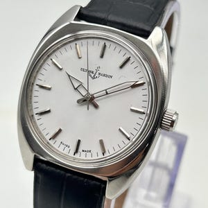 May include: A silver-toned Ulysse Nardin wristwatch with a white face and black leather band. The watch features a rectangular case, silver hands, and a second hand. The watch face displays the brand name and the words "Swiss Made".