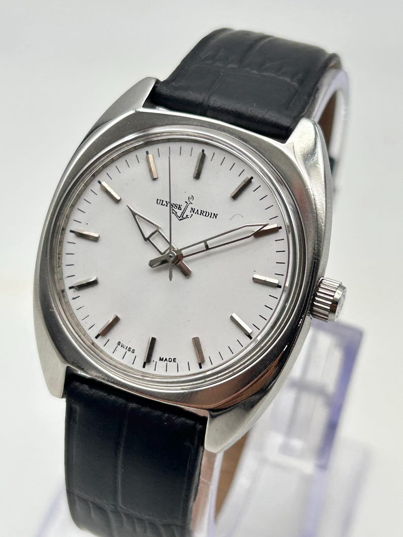 May include: A silver-toned Ulysse Nardin wristwatch with a white face and black leather band. The watch features a rectangular case, silver hands, and a second hand. The watch face displays the brand name and the words "Swiss Made".