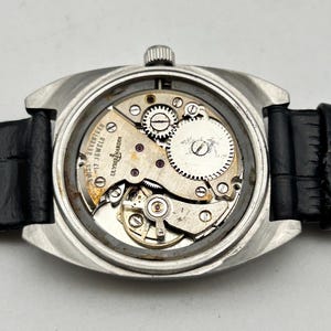 May include: A silver-toned wristwatch with a black leather band, showcasing the intricate internal mechanisms. The watch's back is open, revealing gears, springs, and the inscription "SWISS SEVENTEEN JEWELS ULYSSE NARDIN".