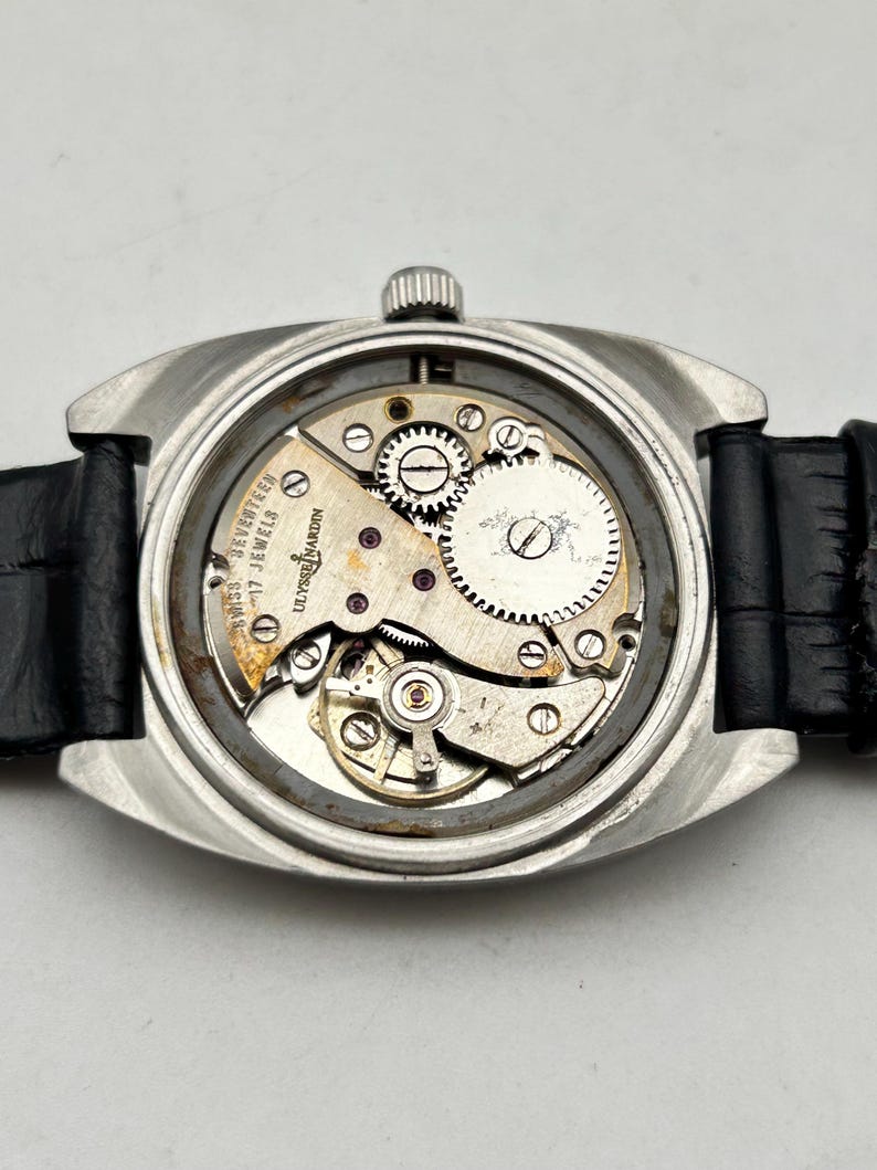 May include: A silver-toned wristwatch with a black leather band, showcasing the intricate internal mechanisms. The watch's back is open, revealing gears, springs, and the inscription "SWISS SEVENTEEN JEWELS ULYSSE NARDIN".