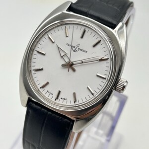 May include: A silver-toned Ulysse Nardin wristwatch with a white face and black leather band. The watch has a rectangular case and a classic design, with the brand name and "SWISS MADE" visible on the face.