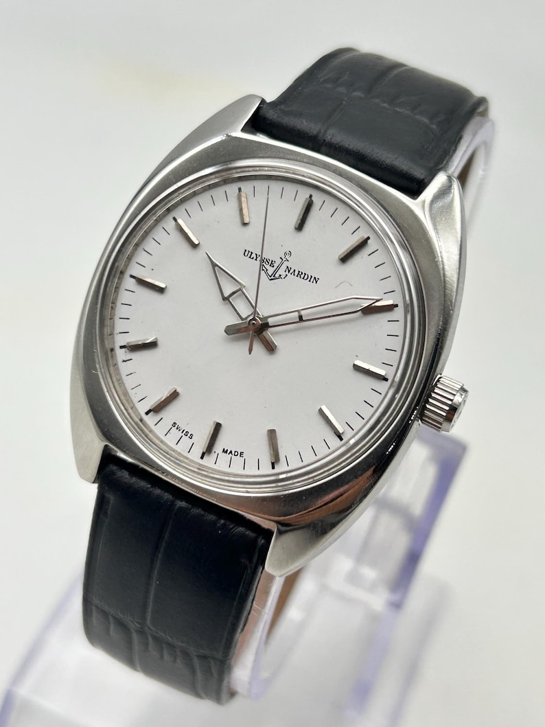 May include: A silver-toned Ulysse Nardin wristwatch with a white face and black leather band. The watch has a rectangular case and a classic design, with the brand name and "SWISS MADE" visible on the face.