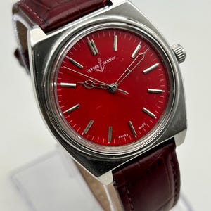 May include: A silver-tone wristwatch with a vibrant red dial featuring the Ulysse Nardin logo and anchor symbol. The watch has silver hour markers, silver hands, and a dark red leather strap with a textured, embossed crocodile-like pattern.