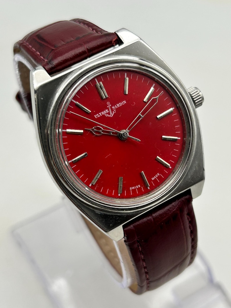 May include: A silver-tone wristwatch with a vibrant red dial featuring the Ulysse Nardin logo and anchor symbol. The watch has silver hour markers, silver hands, and a dark red leather strap with a textured, embossed crocodile-like pattern.
