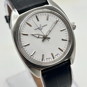 May include: A silver-toned Ulysse Nardin wristwatch with a white face and black leather band. The watch features a round face with silver hour markers and hands. The band is textured and has a buckle closure. The watch is displayed on a clear stand.