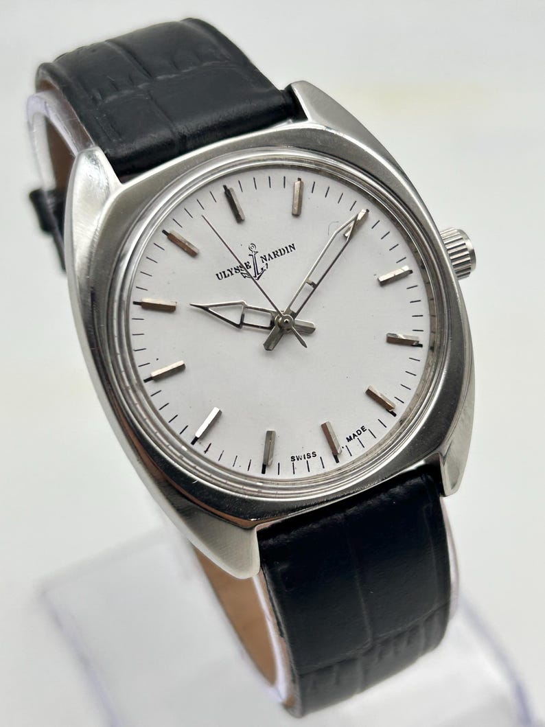May include: A silver-toned Ulysse Nardin wristwatch with a white face and black leather band. The watch features a round face with silver hour markers and hands. The band is textured and has a buckle closure. The watch is displayed on a clear stand.