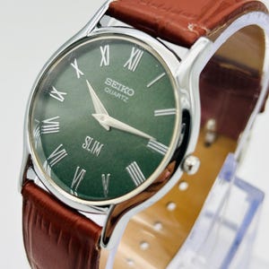 May include: A Seiko Quartz watch with a green face and silver hands and markers. The watch has a silver case and a brown leather band. The word "SLIM" is printed on the watch face. The watch band is approximately 8 inches long.
