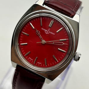 May include: A Ulysse Nardin wristwatch featuring a vibrant red dial with silver-toned hour markers and hands. The watch has a polished silver-tone stainless steel case and a dark burgundy leather strap with a textured, embossed finish.