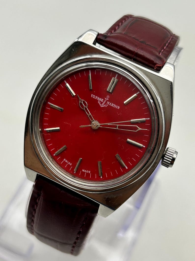 May include: A Ulysse Nardin wristwatch featuring a vibrant red dial with silver-toned hour markers and hands. The watch has a polished silver-tone stainless steel case and a dark burgundy leather strap with a textured, embossed finish.