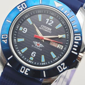 Op de afbeelding: Een Citizen automatisch horloge met een blauwe lunette en band. De wijzerplaat is zwart met witte en blauwe markeringen, en de dag en datum worden weergegeven. De kast is van roestvrij staal en de band van blauw rubber.