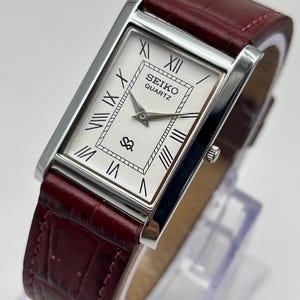 May include: A rectangular Seiko Quartz watch with a silver-tone case and a white face featuring black Roman numerals. The watch has a burgundy leather band. The watch face displays the Seiko Quartz logo.