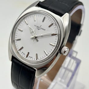 May include: A silver-toned Ulysse Nardin wristwatch with a white face and black leather band. The watch has a rectangular case and a classic design. The watch face features silver hands and markers. The band is textured.
