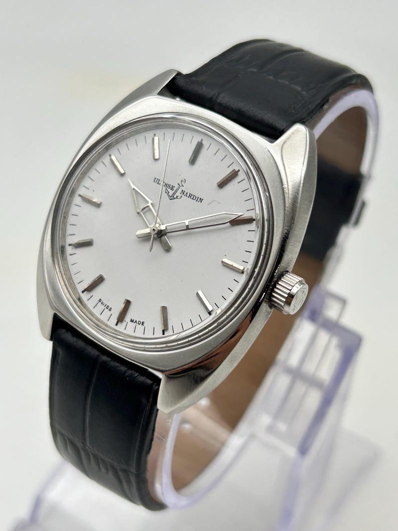 May include: A silver-toned Ulysse Nardin wristwatch with a white face and black leather band. The watch has a rectangular case and a classic design. The watch face features silver hands and markers. The band is textured.