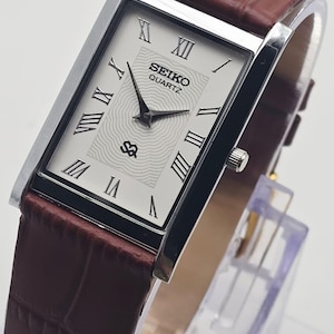 Könnte beinhalten: Eine rechteckige Seiko Quartz-Uhr mit silberfarbenem Gehäuse und weißem Zifferblatt mit schwarzen römischen Ziffern. Das Armband ist aus braunem Leder mit Krokodilmuster. Die Uhr wird auf einem transparenten Ständer präsentiert.