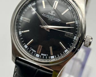 May include: A stainless steel Jaeger-LeCoultre Club watch with a black dial and silver hands and markers. The watch has a black leather band with white stitching. The watch face has the text "JAEGER-LECOULTRE CLUB" and "SWISS MADE T".