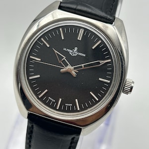 Könnte beinhalten: Eine silberfarbene Ulysse Nardin Uhr mit schwarzem Zifferblatt und schwarzem Lederarmband. Das Zifferblatt hat silberfarbene Zeiger und Markierungen. Die Wörter "Ulysse Nardin" und "Swiss Made" sind auf dem Zifferblatt zu sehen.